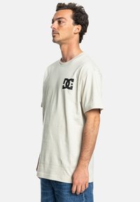 Hellbeige T-Shirt mit kurzen Ärmeln aus Baumwolle, das ein schwarzes DC-Logo auf der linken Brust trägt. Lässiger Schnitt mit Rundhalsausschnitt und weicher Textur.