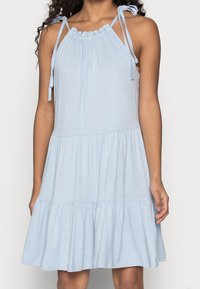 Hellblaustes, ärmelloses Kleid mit gestuften Rock und verstellbaren Bindeträgern, getragen von einer Person mit schulterlangen Locken.