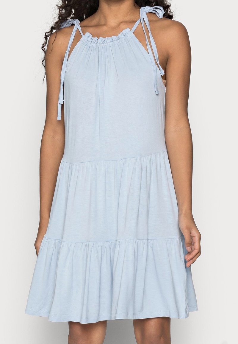 Hellblaustes, ärmelloses Kleid mit gestuften Rock und verstellbaren Bindeträgern, getragen von einer Person mit schulterlangen Locken.