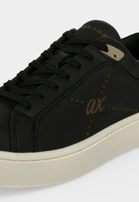 Μαύρο sneaker χαμηλού τύπου με μαύρα κορδόνια, μπεζ λεπτομέρεια στις τρύπες για κορδόνια, λευκή σόλα και χρυσό λογότυπο "ax" με γύρω τελείες στην πλαϊνή επιφάνεια.