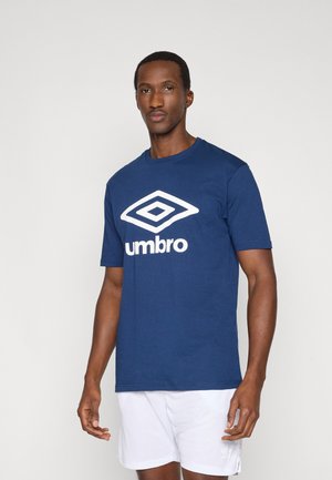 Hombre con camiseta azul marino de Umbro y pantalones cortos blancos, de pie frente a un fondo claro y liso, mirando hacia adelante con expresión neutral.