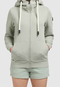 Donna che indossa una felpa leggera verde mela con zip e lacci bianchi, abbinata a pantaloni corti, con le mani nelle tasche, su uno sfondo chiaro e semplice.