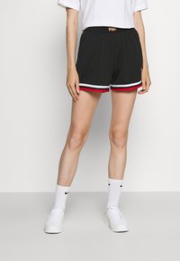 Henkilö, joka on pukeutunut mustiin verkkokankaiseen urheilushortsit, joissa on punainen ja valkoinen reunus, valkoisiin yksivärisiin sukkiin, valkoisiin lenkkariin ja valkoiseen T-paitaan, seisoo tasaisen seinän vieressä.