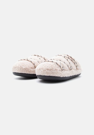 Chaussons beiges duveteux avec une surface extérieure en peluche, une semelle en caoutchouc noire et des lanières croisées en couleur contrastante pour une texture supplémentaire.