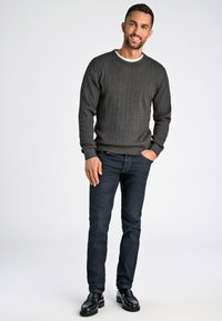 Tummanharmaa palmikkoneule, valkoinen kaulus, yhdistettynä tummaan farkkuihin ja mustiin loafer-kengiin. Malli seisoo rentona yksi käsi taskussa.