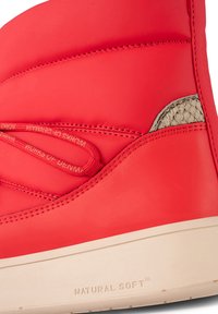 Botín de tobillo sintético rojo con textura suave, cordones elásticos y suela natural suave. Presenta un detalle tejido cerca del tobillo.