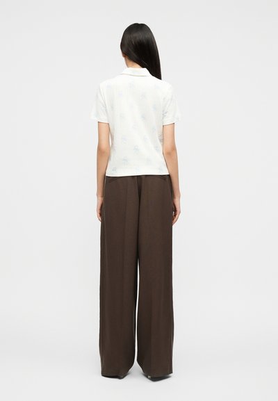Femme debout, de dos, portant une chemise blanche à manches courtes avec col et un pantalon large marron, sur un fond uni.