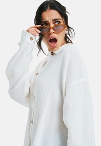 Witte, gestructureerde button-up shirt met lange mouwen. Model draagt kleine, ovale bruine zonnebril en kijkt verrast met een open mond.