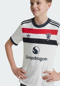 Maillot de sport blanc avec des rayures horizontales rouges et noires, arborant le logo Adidas, la marque Snapdragon et trois rayures noires sur les épaules.