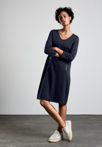Robe bleu marine à mi-longueur avec un col rond, des manches longues et des poches zippées à l'avant. Le tissu semble doux et lisse. portée avec des sneakers légers.