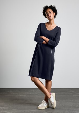 Robe bleu marine à mi-longueur avec un col rond, des manches longues et des poches zippées à l'avant. Le tissu semble doux et lisse. portée avec des sneakers légers.
