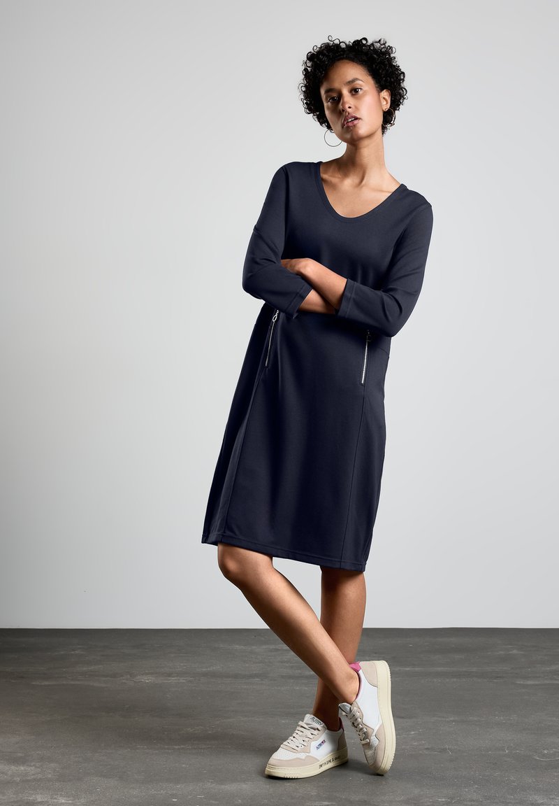 Robe bleu marine à mi-longueur avec un col rond, des manches longues et des poches zippées à l'avant. Le tissu semble doux et lisse. portée avec des sneakers légers.