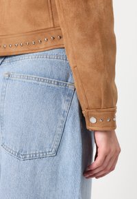 Brun semsket skinnjakke med sølvnagler langs kanten og mansjettene, paired med lyseblå denim bukser med baklomme.
