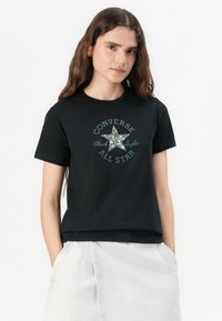 Musta puuvillainen T-paita, jossa on graafinen tähti -kuvio ja teksti "Converse Chuck Taylor All Star" pyöristettynä. Lyhythihaiset.