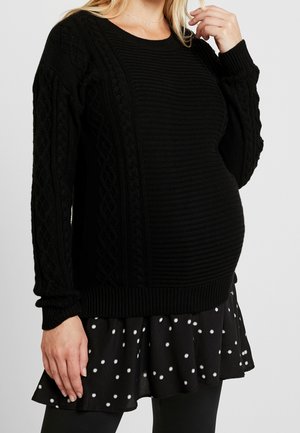 Gravid kvinde iført en sort tekstureret strikket sweater med kabelmønstre og et sort skørt med hvide polkaprikker.