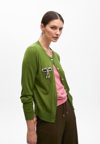 Grön kavaj med en framknäppning, prydd med en dekorativ brosch, lagd över en rosa topp och matchad med bruna byxor. Slät textur.