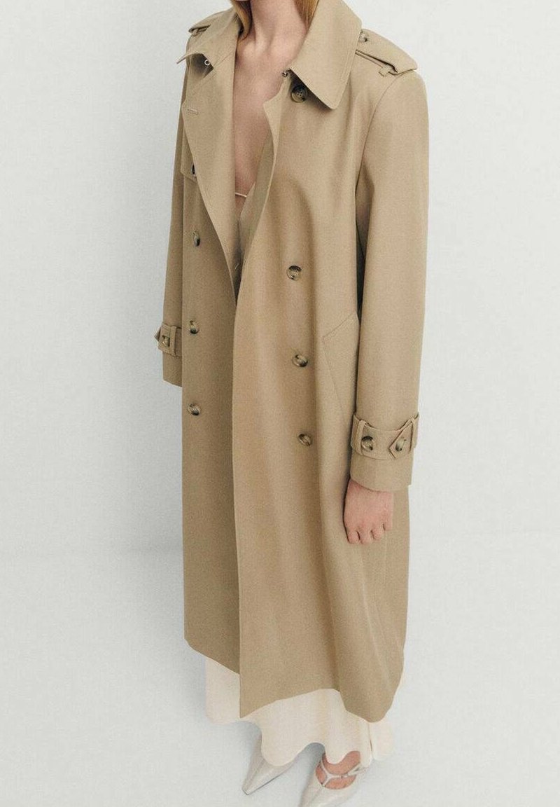 Personne portant un long trench-coat beige sur une robe blanche et des chaussures blanches pointues sur un fond clair uni.