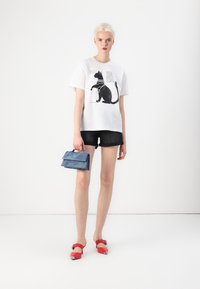 Hvit t-skjorte med svart katt grafikk, parret med mørke denimshorts. Holder en blå brettet veske, og har på seg røde sko med sølvdetaljer.