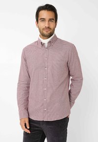 Rood en wit geblokt overhemd met kraag, voorzien van een linkse borstzak en bruine knopen. Draagt over een witte turtleneck.