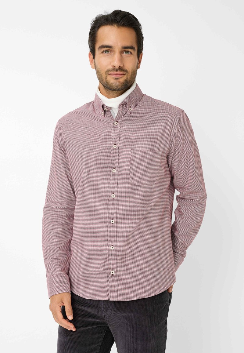 Rood en wit geblokt overhemd met kraag, voorzien van een linkse borstzak en bruine knopen. Draagt over een witte turtleneck.