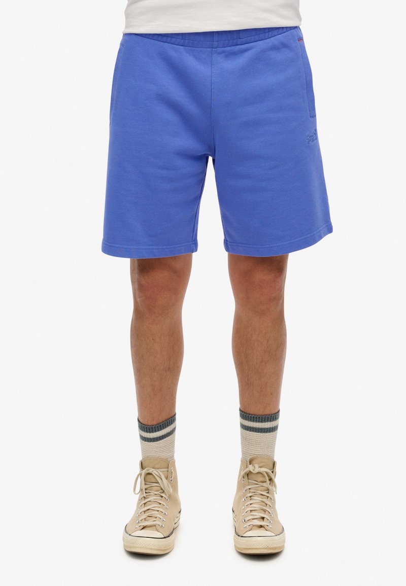 Blauwe katoenen shorts met een elastische tailleband, rechte pasvorm en zijzakken. Draagt een paar beige hoge sneakers en gestreepte sokken.