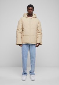 Beige pufferjacka med huva, med quiltat mönster och framdragkedja. Matchat med ljusa denimjeans och vita sneakers.