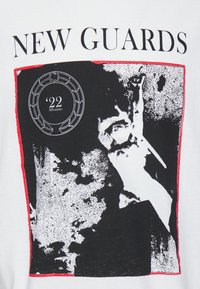 Bílé tričko s černobílým grafickým designem a červenými akcenty, na kterém je vytištěn text "NEW GUARDS" a kruhové logo s "22."