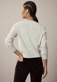 Šviesiai pilkas, margas megztinis su ilgomis rankovėmis, apvaliu iškirpimu ir raštuotais rankogaliais. Sugrupuotas su juodomis faktūrinėmis kelnėmis, rodančiomis nugarinę pusę.