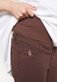 Os jeans de denim castanho apresentam um design ajustado, detalhe bordado de folha no bolso e uma textura suave. Uma blusa branca cobre parcialmente os jeans.