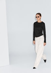 Sort langærmet top, hvide brede bukser og sorte spidse sko. Modellen bærer mørke solbriller. Minimalistisk baggrund.