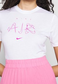 Bílý bavlněný tričko s růžovým textem a logem "Nike AJ". V páru s růžovou skladanou sukní, která má hladkou texturu a elastický pas.