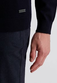 Marineblauer gestrickter Pullover mit gerippten Bündchen, versehen mit einem kleinen schwarzen Etikett mit dem Markennamen am Saum, getragen mit strukturierten dunklen Hosen.