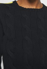 Svart stickad tröja med ett kabelmönster, rund halsringning och en ljusgul detalj på axeln. Tjockt material och texturerad.