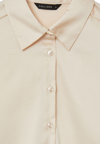 Blusa beige con colletto classico, dotata di sei bottoni frontali, texture liscia e dettagli di cucitura minimali. Etichetta del marchio: Calliope.
