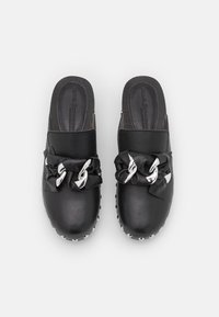 Sabots en cuir noir avec un nœud en satin agrémenté de chaînes argentées et d'une semelle avec clous. Texture lisse, design à bout fermé, style à enfiler.