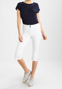 Kvinna som bär en marinblå topp med kort ärm, vita högmidjade capribyxor och vita sneakers, stående mot en enkel bakgrund.