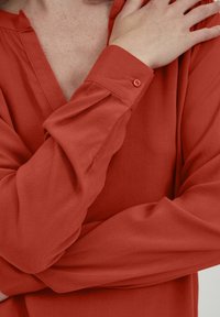Blusa rosso ruggine con scollo a V e maniche lunghe. Tessuto liscio, dettaglio con bottoni al polsino e texture sottile visibile.