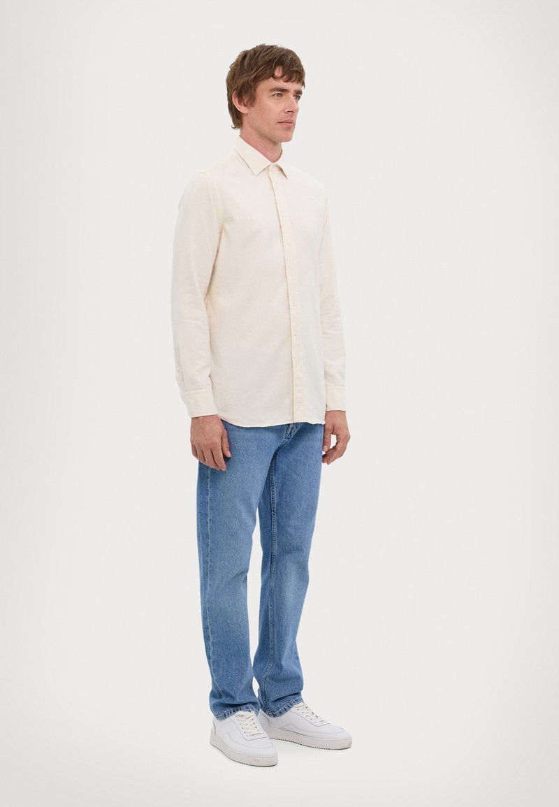 Camisa de manga larga beige claro con un diseño de botones, combinada con jeans azul claro y zapatillas blancas, de pie contra un fondo liso.