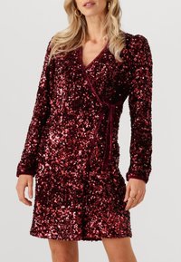 Robe bordeaux à sequins avec manches longues, col en V et taille à nouer. Caractérisée par une texture scintillante et un design portefeuille.