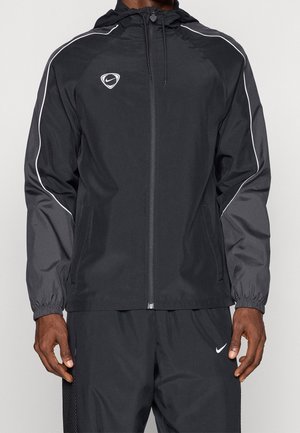 Homme portant une veste à capuche zippée noire avec des manches grises et un liseré blanc, associée à un pantalon noir Nike avec logo swoosh blanc.