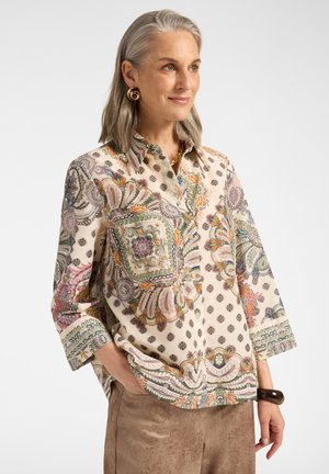 Frau mit grauen Haaren, die eine beige Bluse mit Paisley-Muster, braune Wildlederhose, goldene Ohrringe und ein dunkles Armband trägt und mit einer Hand in der Tasche steht.