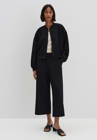 Veste en cuir noire avec col rond, boutons sur le devant et poignets élastiques. Associée à un pantalon noir à jambes larges et des sandales plates noires.