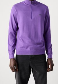 Man i lila långärmad tröja med en fjärdedels dragkedja och svarta byxor, stående mot en enkel bakgrund.