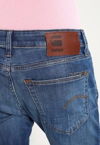 Blå denimjeans med en läderlogotyp på baksidan, med stickade detaljer och två bakfickor med kontrasterande tråd.