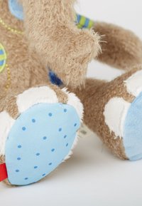 Pattes douces d'ours en peluche marron avec des semelles en tissu bleu ornées de petits points bleus, des accents de fourrure blanche et une partie d'un patch coloré sur le corps.