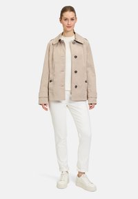 Beige Baumwollmischung Jacke mit kragenförmigem Design, ausgestattet mit sechs dunklen Knöpfen und hochgerollten Ärmeln, getragen über einem weißen Pullover und weißen Hosen.