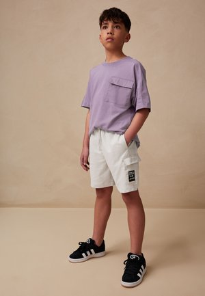 Kortærmet lavendelfarvet t-shirt med brystlomme, kombineret med hvide cargoshorts med sidelomme og sort mærke, samt sorte sneakers.
