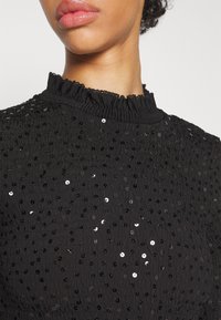 Femme portant un haut noir à col montant avec de petites paillettes noires et argentées sur un tissu texturé, montrée du menton à la poitrine.