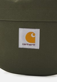 Sac en tissu vert avec une texture lisse. Présente une étiquette logo Carhartt blanche avec une icône orange et un texte bordeaux.