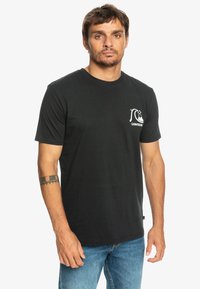 Black cotton T-shirt with short sleeves, crew neck, and a white logo on the left chest. Worn with blue denim jeans.